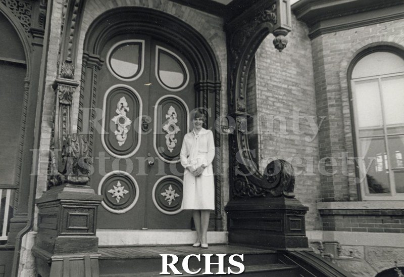 Black and white photograph of Judy Berg standing in front of ornate doors of the Berg house, a brick house.