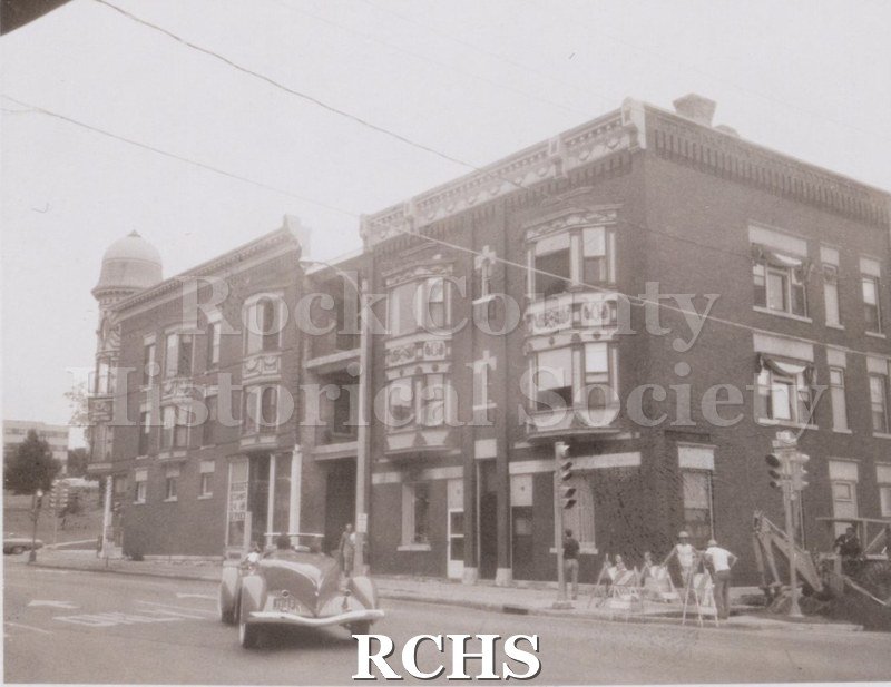 Black and white photograph of the three story apartment building at 52 E.Court St. There are workmen working on the sidewalk and a convertible on the street.