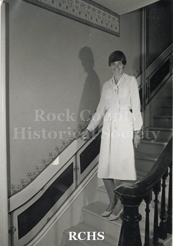 Black and white photograph of the Berg residence. Shows a woman standing on a staircase. Note the stenciling on the wall.