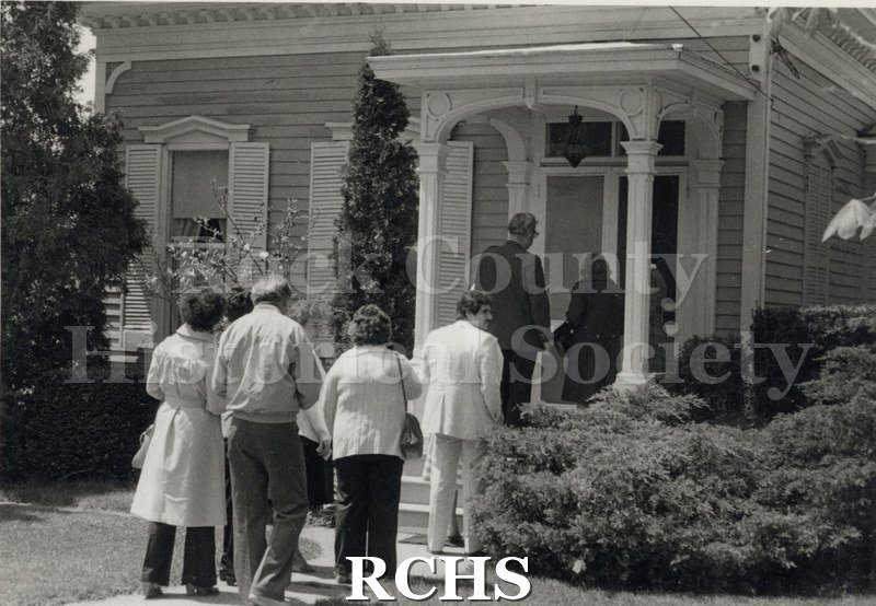 Black and white photograph of the Howarth house. Shows several people standing on the front porch of a two story house waiting for the tour.