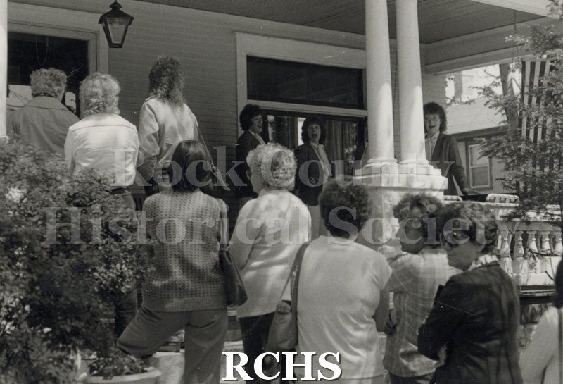 Black and white photograph of four unidentified women standing on a porch, singing, while people are listening.