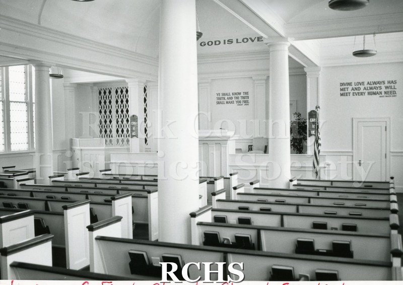 Black and white photograph of the interior of First Church of Christ Scientist. Shows the pews and a sign: "God is love" on the wall.