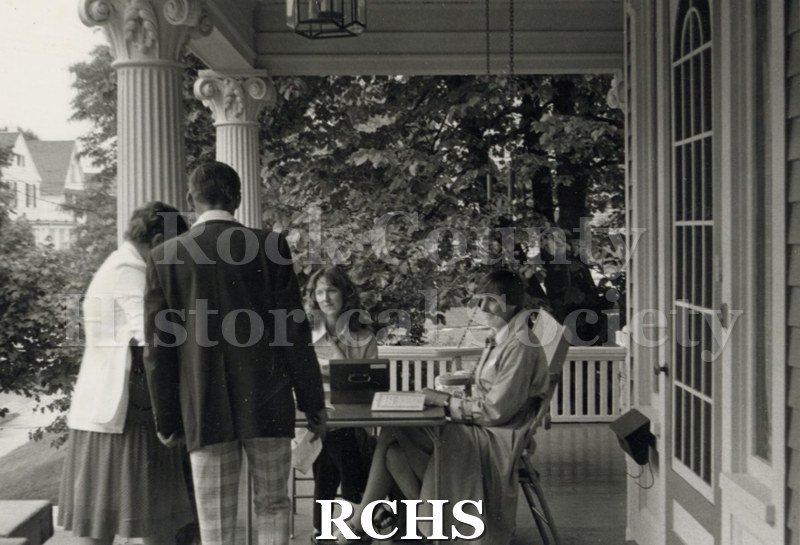 Black and white photograph of Judy Berg and Lynn Welscher sitting at a card table on the porch of a house with a couple ready to pay for admission.