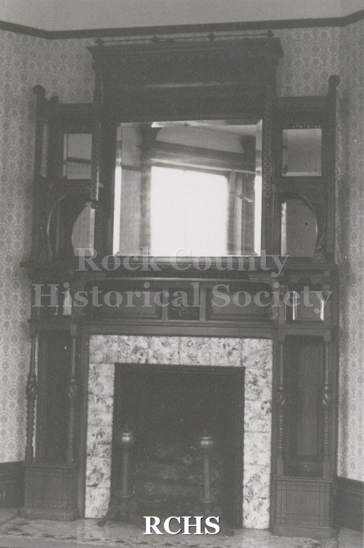Black and white photograph of a fireplace with a large mirror above it and andirons on the floor of Lynn Dowdy's home.