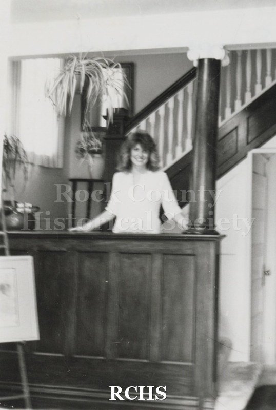 Black and white photograph of an unidentified woman standing behind a section of an entry way with a staircase behind her and a hanging plant above her at the.