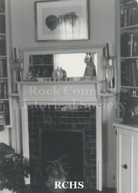 Black and white photograph of a fireplace with a plant in front of it at the Swanson home. There are several objects on the mantel and a mirror above it.