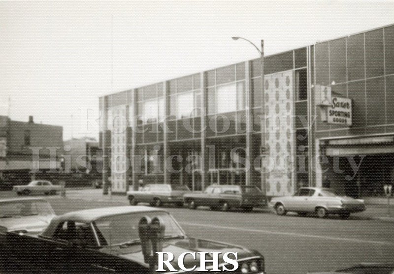 Black and white photograph of the Rock County Bank. Shows a two story building with design panels on the front. Bank is next to Saxer Sporting Goods.