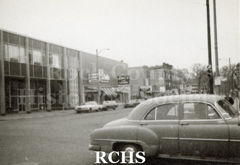 Black and white photograph of N. Main Street. Shows a row of stores on the left side of the photo, including Saxers Sports, W. G. Bullis, Dells Cleaning.
