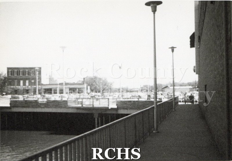 Black and white photograph of the Parking Plaza, Janesville, WI, from the river. Shows the cars parked in the plaza and the walkway into the plaza.