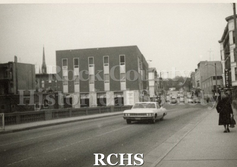 Black and white photograph of downtown Janesville businesses showing the Milwaukee Street bridge with one car passing and a pedestrian on the sidewalk.