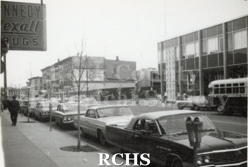 Black and white photograph of downtown Main Street Janesville, showing part of sign for "Kennedy Rexall Drugs" on left side.