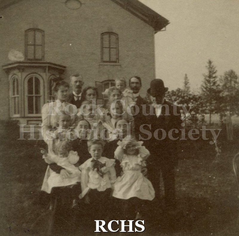 Black and white photograph of 15 adults and children standing in front of the home of Mr. and Mrs. Wm. Menzies on Tarrant Road, Rock County, Wis.