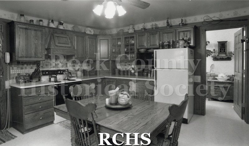 Black and white photograph of a kitchen with a wooden table and four chairs in the middle; wooden cabinets on two sides with the stove and refrigerator in between.