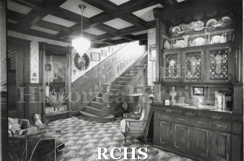 Black and white photograph of residence at 210 S.Jackson. Shows a room with a stairway in the middle of the photo and a breakfront on the right.