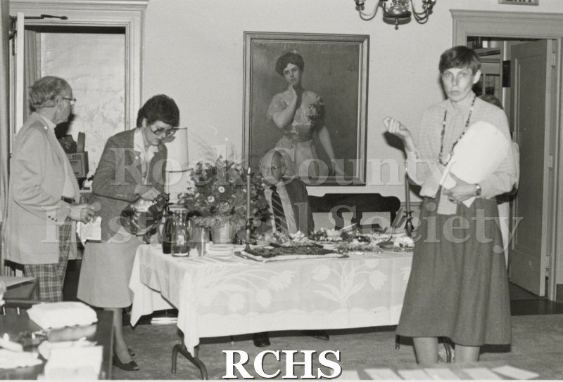 Black and white photograph of four people attending a tea. One man is seated and the other three are standing near the dessert table.