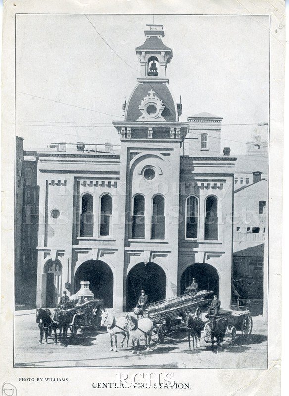 Historic image of Black and white copy print of a two story fire station with a belltower. In front are two pieces of equipment pulled by horses.