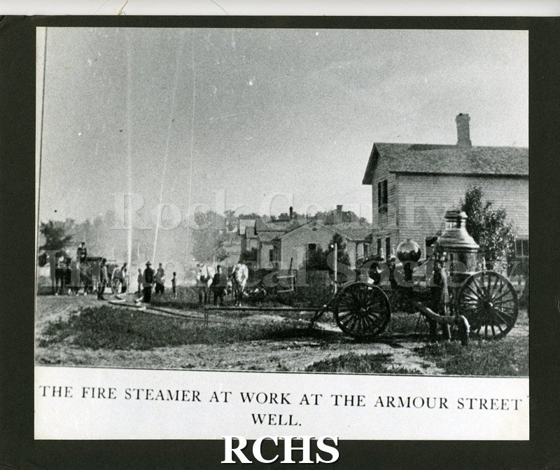 Copy print photograph of an old fire engine. There are several firefighters on the left and another engine pulled by horses.