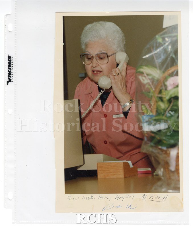 Historic image of Color photogrpah of Mercy Hospital volunteer Katie Hall answering the phone at the front desk. On bottom: "Front Desk-Mercy Hospital-Al Hoch".