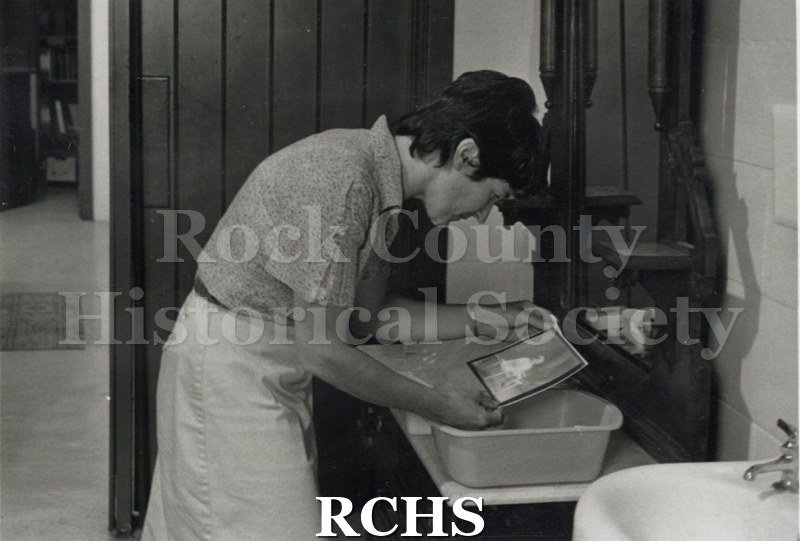 Black and white photograph of Ann Wilmot looking at a picture. There is a sink on the right.