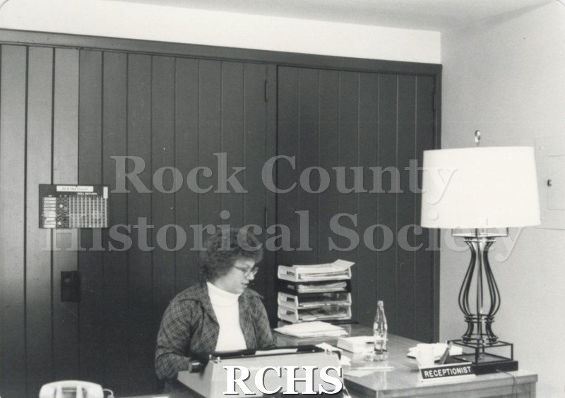 Black and white photograph of Lois Coulthard working on a typewriter. There is a sign: "Receptionist" on the desk beside a lamp.
