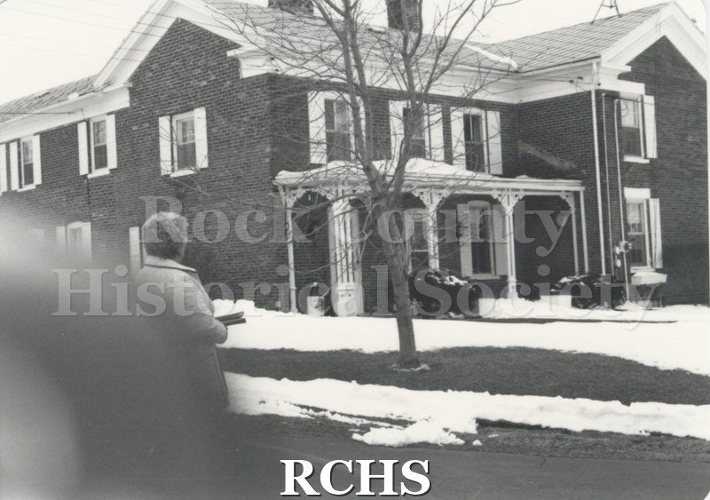 Black and white photograph of Marjorie Stevenson standing near a two story house. There is snow on the ground.