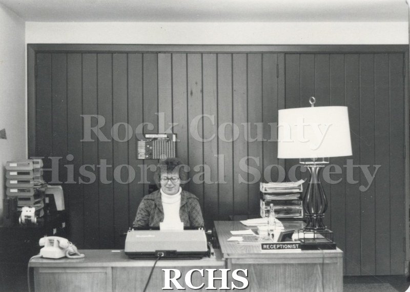 Black and white photograph of Lois Coulthard working on a typewriter. There is a sign: "Receptionist" on the desk beside a lamp.
