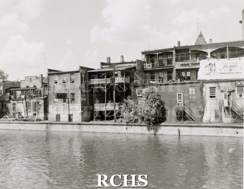 Black and white photograph of 13 N. Main St. Shows the back side of stores on Main Street. The river is in the foreground.
