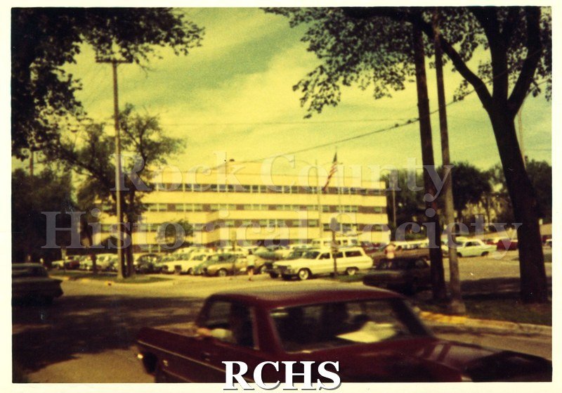 Color photo of the Rock County Courthouse with cars parked in front of it and a flag flying.