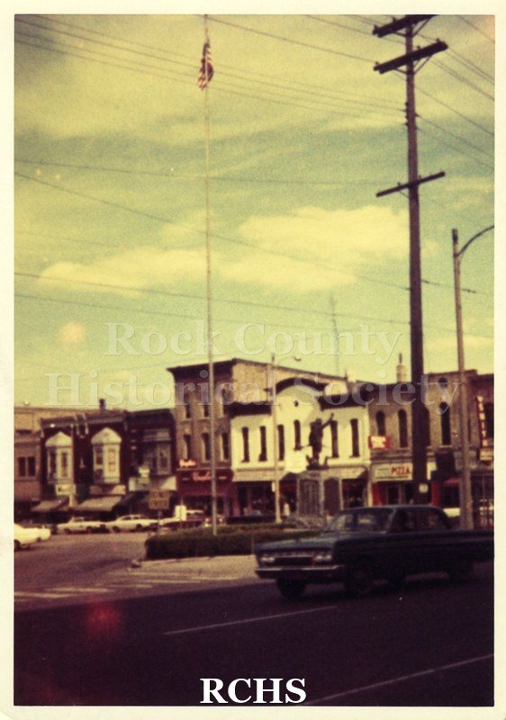 Color photo of Corn Exchange. Shows store fronts from a distance. There is a large flagpole in the front.