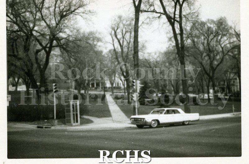 Black and white photograph of Court House Park. Shows a phone booth on the corner by the stop sign and a car stopped at the stop sign.