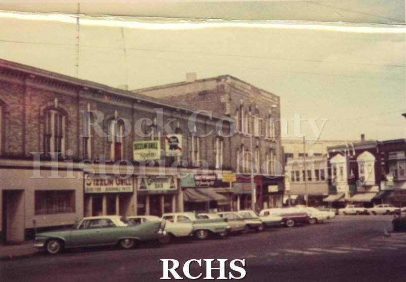 Color photo of downtown Janesville. There are store signs for "Sizzlin' Grill"; "Gunderson's Bar" and others not readable. Area is across from Corn Exchange.