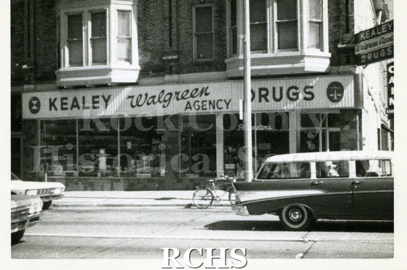 Black and white photograph of Kealy Drugs. Shows a two story building with a sign for "Kealey Walgreen Agency Drugs" visible on storefront.