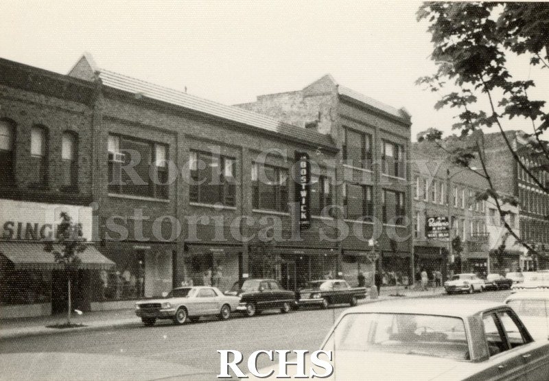 Black and white photograph of South Main Street in downtown Janesville, looking at the builidings with "Singer" and "Bostwick" signs.