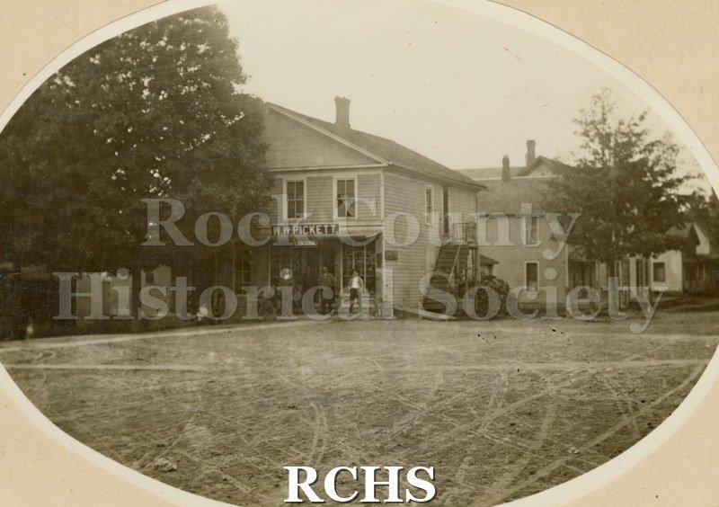 Black and white photograph of the W. W. Pickett Cash Store Janesville, Circa 1903. Shows a two story building with a sign over the porch: ""W. W. Pickett".