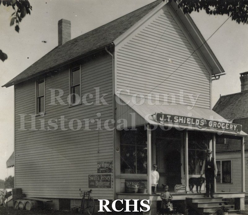 Black and white photograph of the Joseph T. Shield's Grocery. This is a two story store building with a porch on the front. On top of the porch is a sign: "J.T.