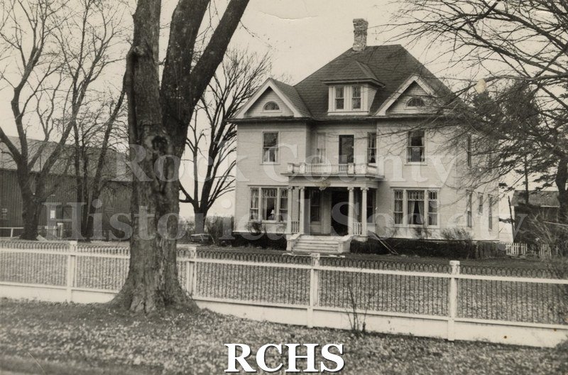 Black and white photograph of Maple Lawn Farm. Shows a three story house with a porch on the front. There is a fence in the foreground.