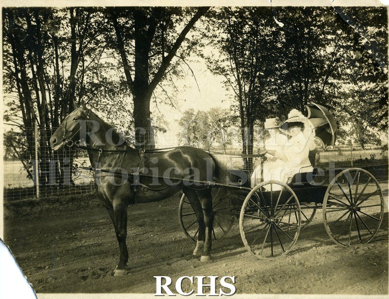 Black and white photograph of Harriet Parsons and Elizabeth Schickey in white dresses in a carriage pulled by a dark horse.