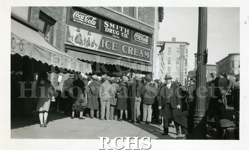 Black and white photograph of a crowd of people are standing in front of Smith Drug Co. Above the awning are signs for Coca-Cola and Ice Cream.