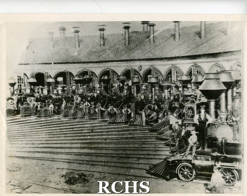Historic image of Black and white copy print of several train engines in front of the round house with men in front of the cowcatchers.