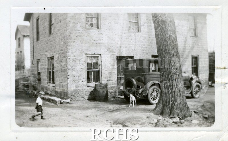 Black and white photograph of a two story house with a car parked in front of it. There is a dog by the car and a small boy walking along the left side.