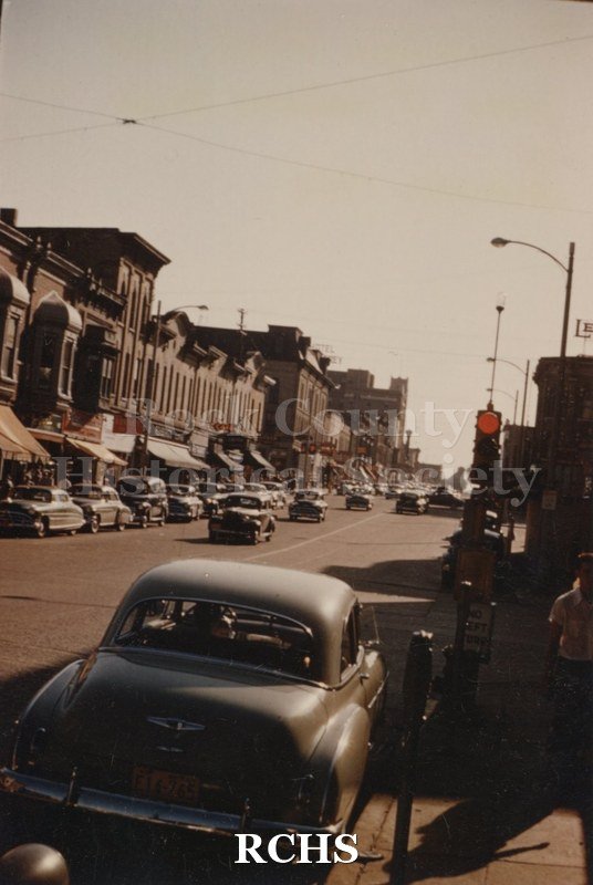 Color photo of Milwaukee Street, Janesville, WI, showing stores along the way and many cars on the street.