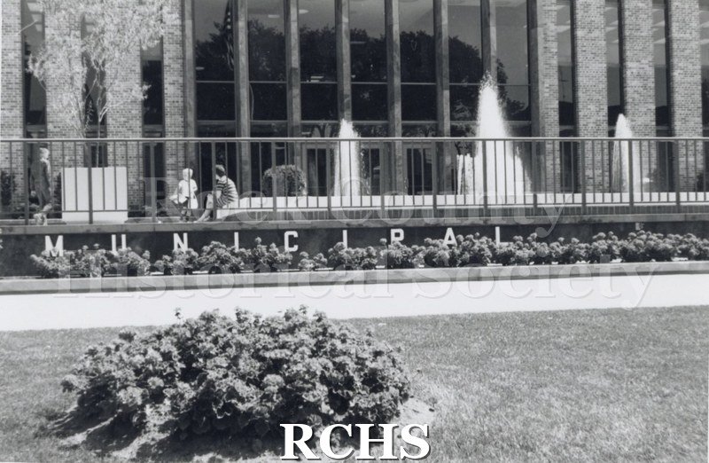 Black and white photograph of Janesville City Hall, showing the front with Municipal spelled out and fountains working. Three people are visible behind the fence.
