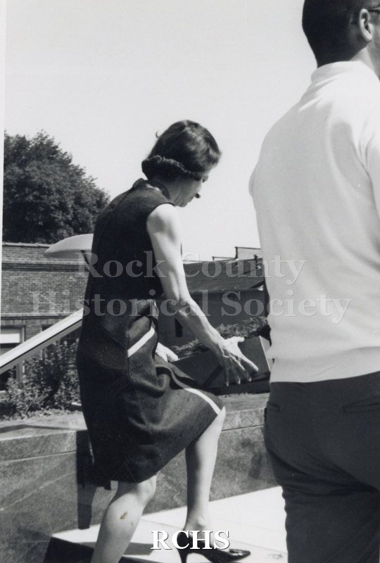 Black and white photograph of unidentified woman walking up a step, carrying a box. She is wearing a dark colored sleeveless dress.