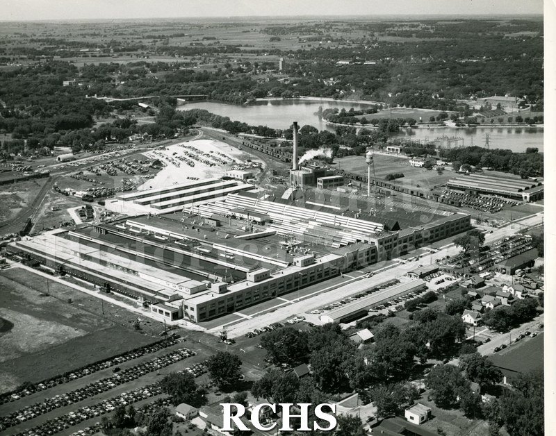 Black and white aerial view of GM plant. Shows plant in middle with cars parked in lots around building and houses in front and behind building.