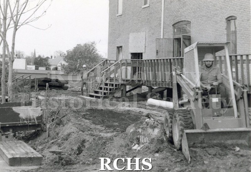 Black and white photograph of a front end loader being operated by a man in a hard hat. Another man is standing in front of the sidewalk in to the building.
