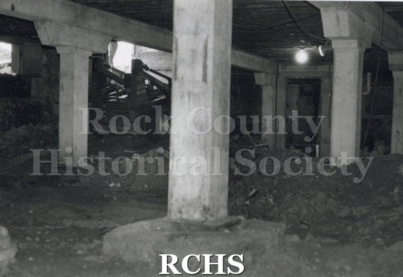 Black and white photograph of a front end loader inside a building. There are two pillars holding up the roof.