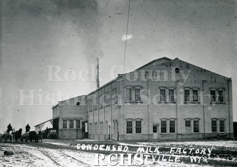 Historic image of Black and white copy print of the Condensed Milk Factory, a two story building with two sets of horse teams on the left side.