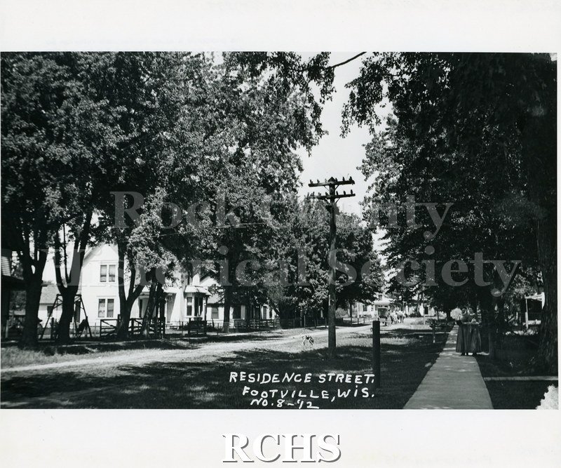 Historic image of Black and white copy print of a street in Footville. There is a horse and buggy down the street and two women strolling on the right sidewalk.