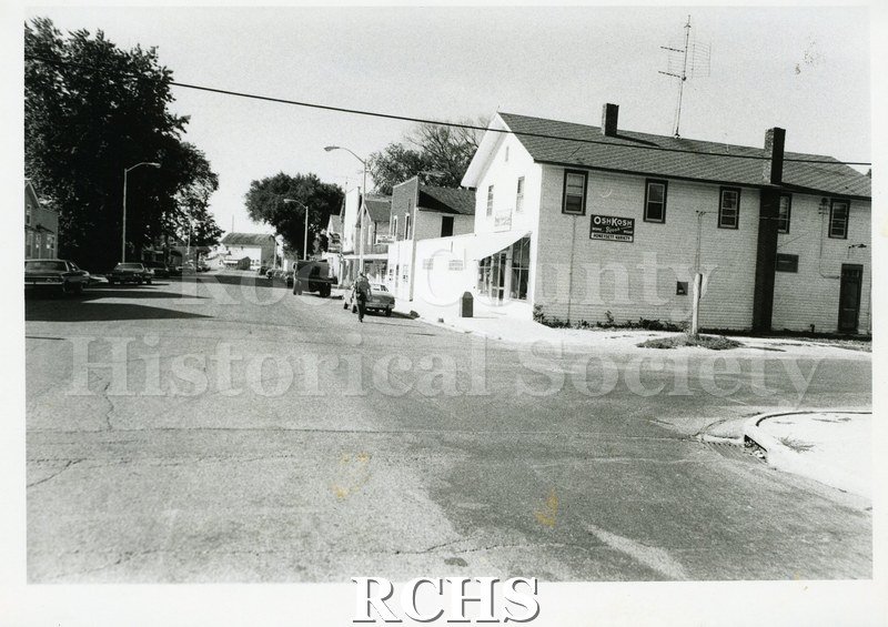 Black and white photograph of an unidentified street in Footville, WI. Sign on the store side reads: "Oshkosh Honeysett Variety".
