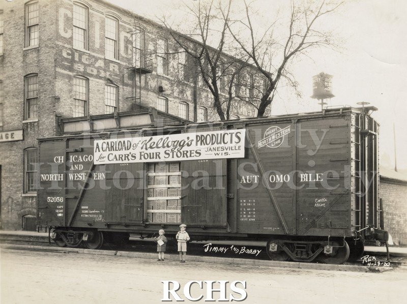Black and white photograph of a railroad car with the sign: "A carload of Kellogg's products for Carr's four stores Janesville, Wis." on the side.